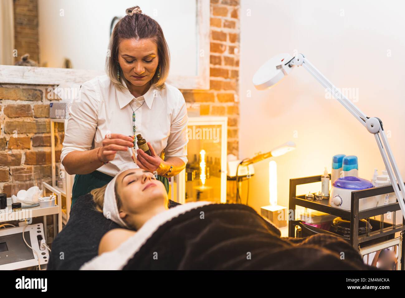 Kosmetikerin, die ein paar Tropfen öliges Serum auf das Gesicht ihrer Klientin aufträgt. SPA-Tageskonzept. Innenausstattung des Hautpflegesalons. Hochwertiges Foto Stockfoto