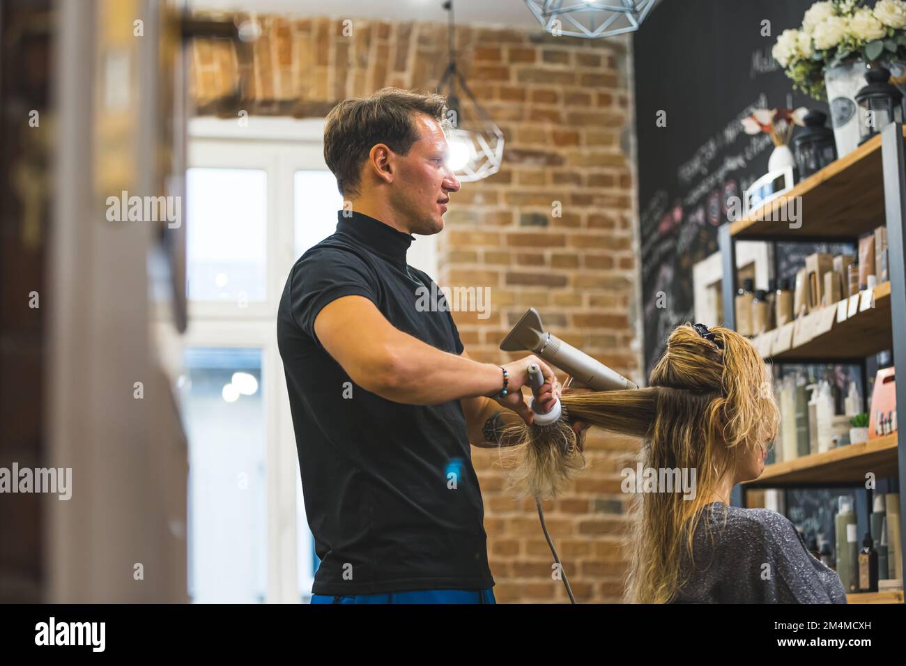 Ein männlicher Friseur, der lange blonde Haare seines Klienten trocknet. Friseurinnenraum. Speicherplatz kopieren. Hochwertiges Foto Stockfoto