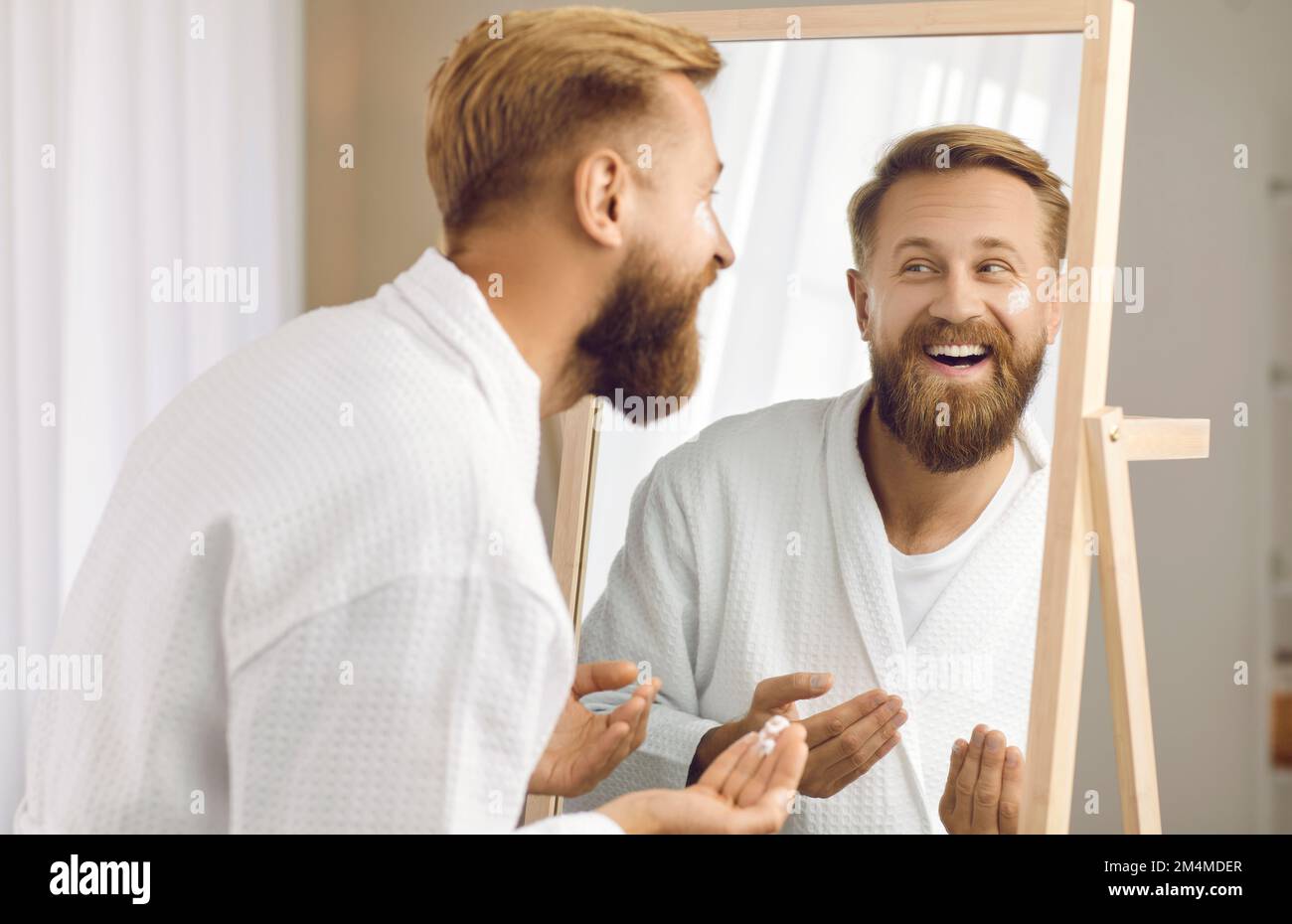 Ein lustiger Mann, der vor dem Spiegel steht, trägt fröhlich Kosmetikcreme auf sein Gesicht auf. Stockfoto
