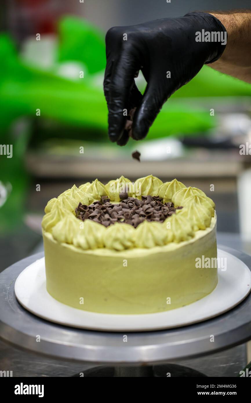 Der Konditoreidesigner streut den grünen, mattierten Schokoladenkuchen mit kleinen pistacchio- und dunklen Schokoladenstücken Stockfoto
