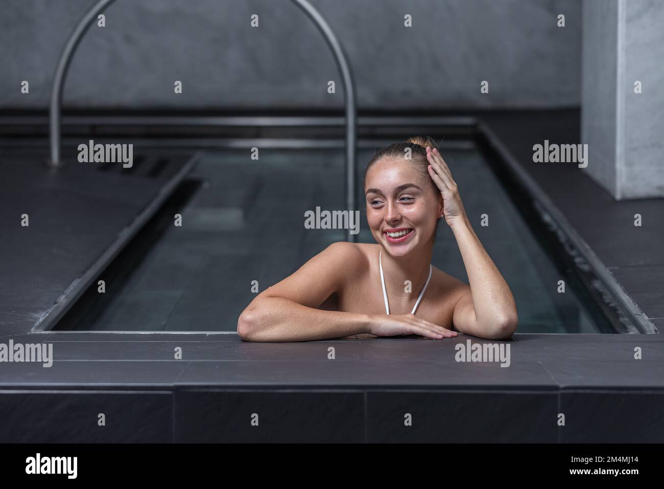 Glückliche junge Frau, die mit einem Lächeln wegsieht und nasses Haar berührt, während sie sich tagsüber im sauberen Pool im Spa-Resort ausruht Stockfoto