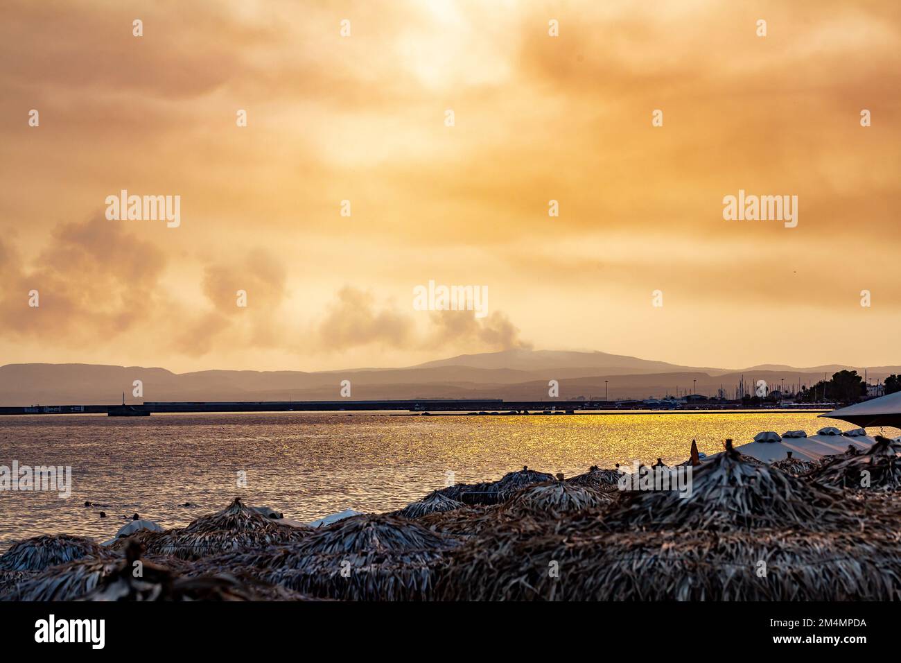 Ruhige Küstenküste mit Bergpanorama und Feuer am abgelegenen Horizont. Orangefarbene Wolkenlandschaft in tropischer Lage im Freien Stockfoto