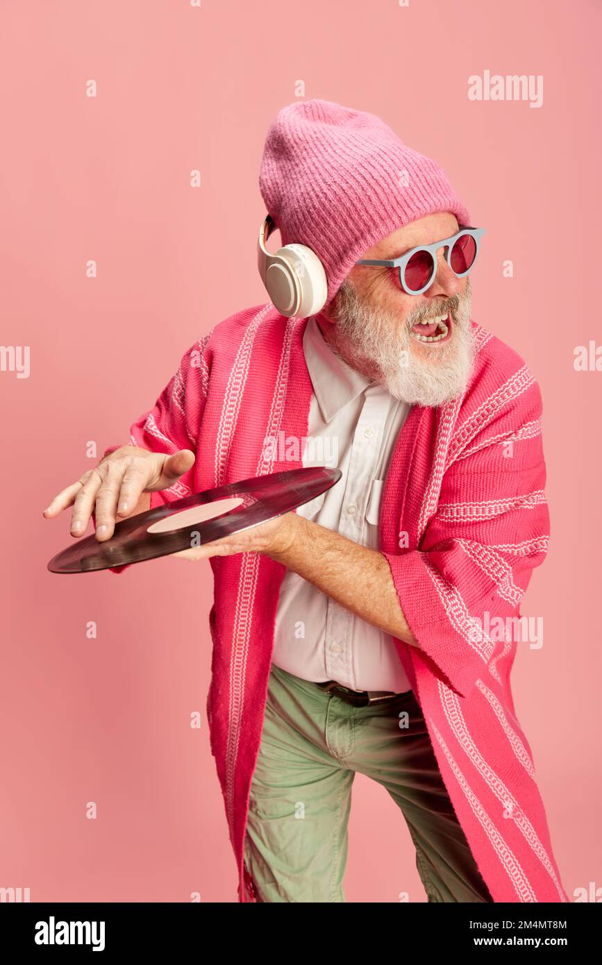 Gefühlvoller alter Mann in elegantem, hellen Outfit mit Strickmütze, Sonnenbrille, Kopfhörern und Vinyl auf pinkfarbenem Hintergrund. Dj Stockfoto