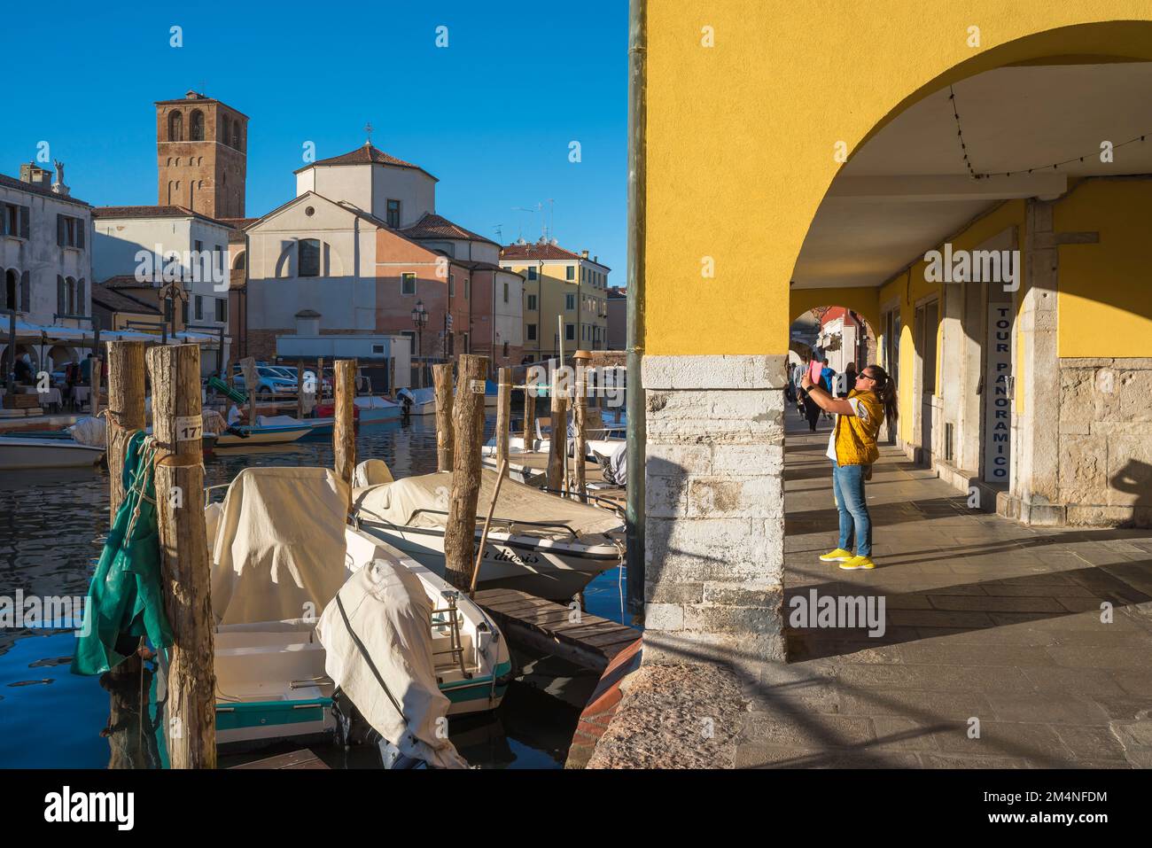 Reisefotografie Frau, Blick auf eine Frau, die in einer farbenfrohen Kolonnade in Chioggia steht und ein Foto von einem malerischen Kanal macht, Comune of Venice, Italien Stockfoto