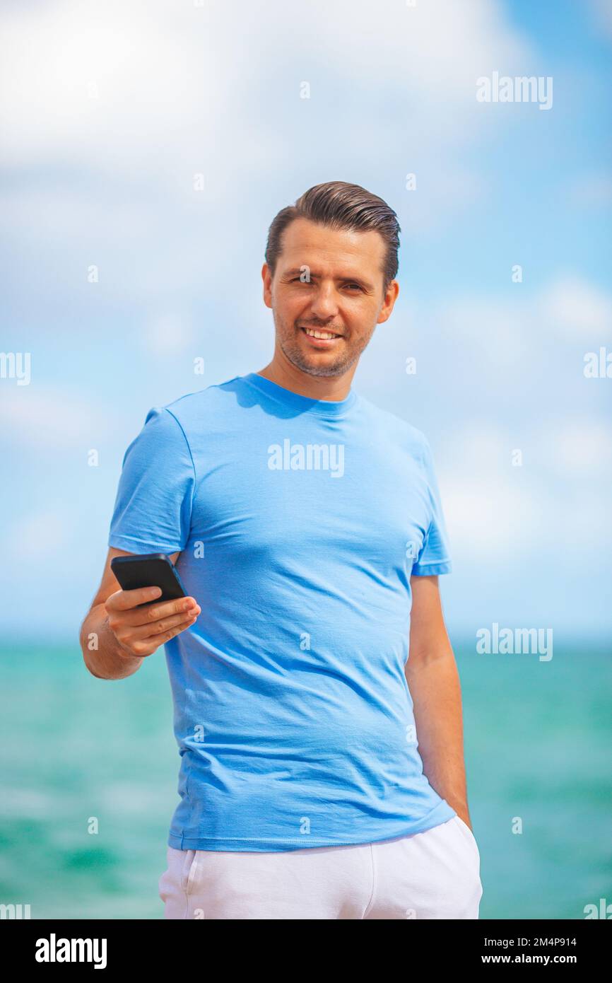 Junger Mann, der sein Handy in der Hand hält, während er am Strand spaziert Stockfoto