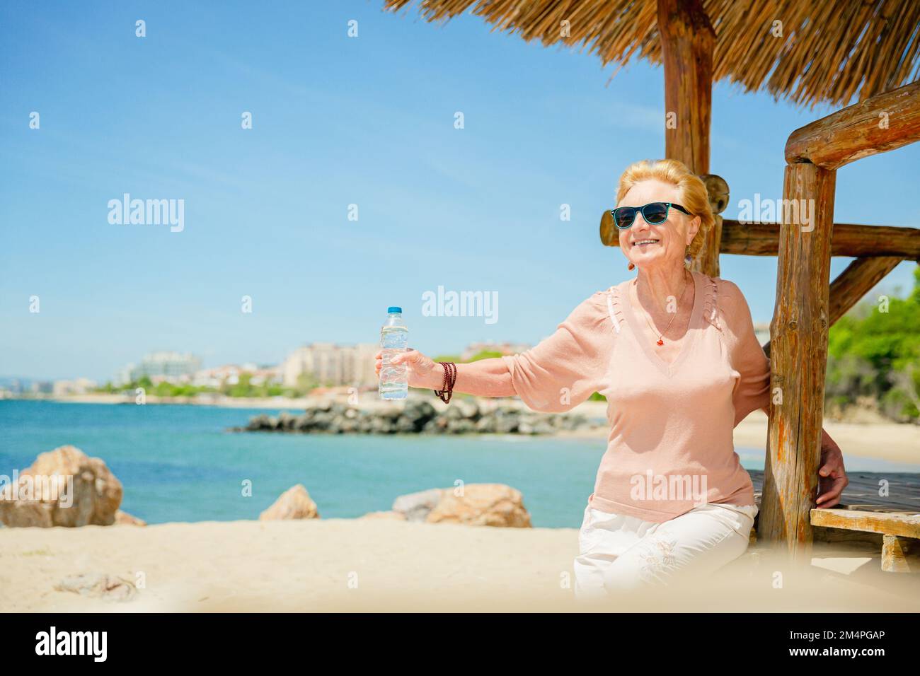 Ältere Frau, die mit einer Flasche Wasser am Strand sitzt Stockfoto