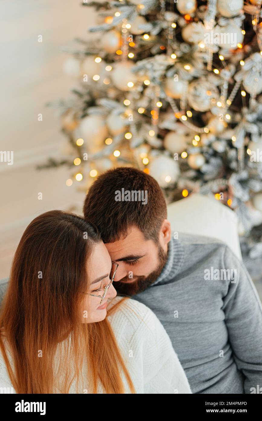 Ein verliebtes Paar, das sich anlächelt, auf dem Sofa neben Weihnachten sitzt, Neujahrsbaum Stockfoto