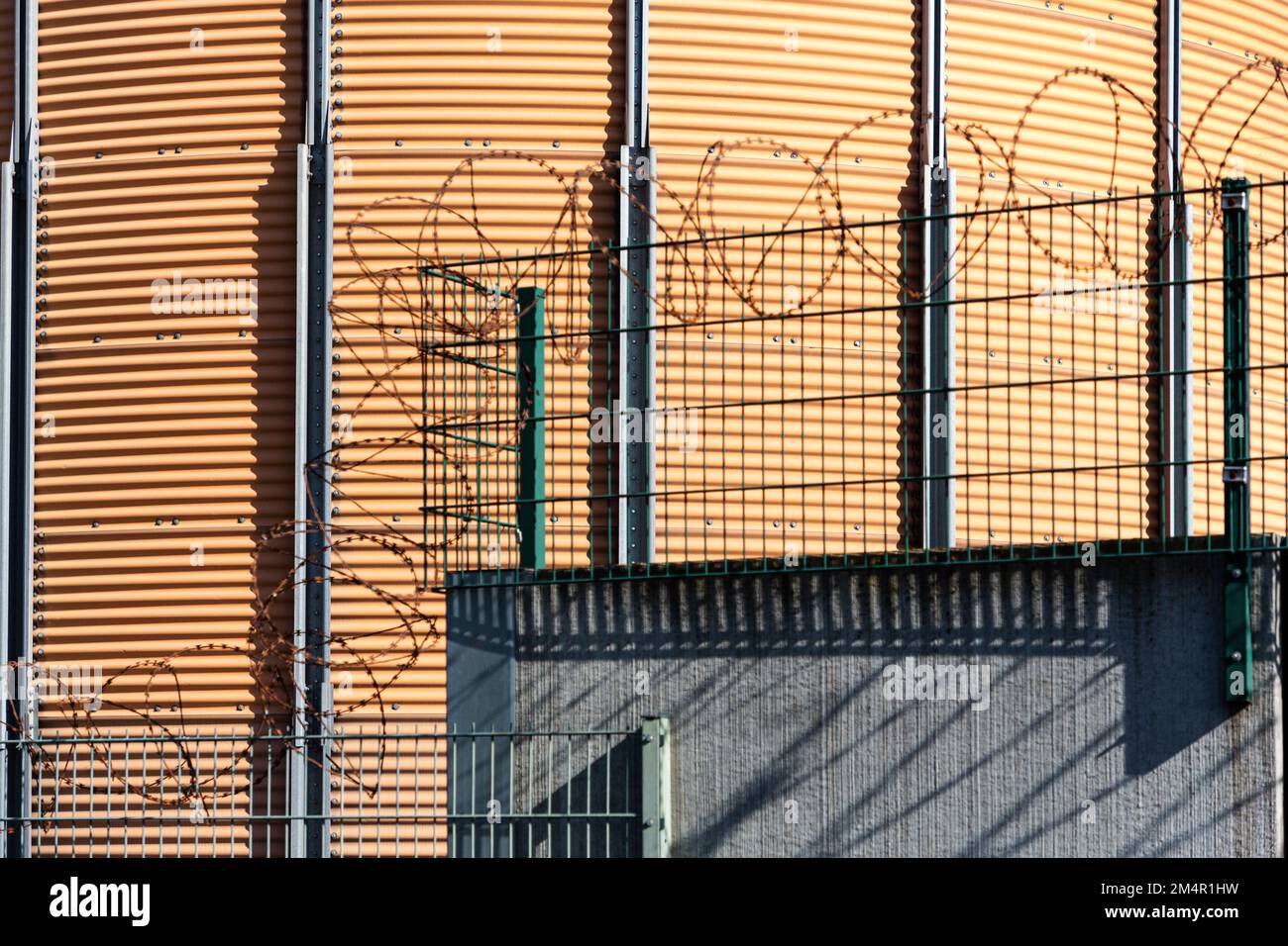 An einer Wand befindet sich ein Zaun, an dem Stacheldraht angebracht wurde. Im Hintergrund befindet sich eine runde orangefarbene Wand eines Industriebaums. Stockfoto