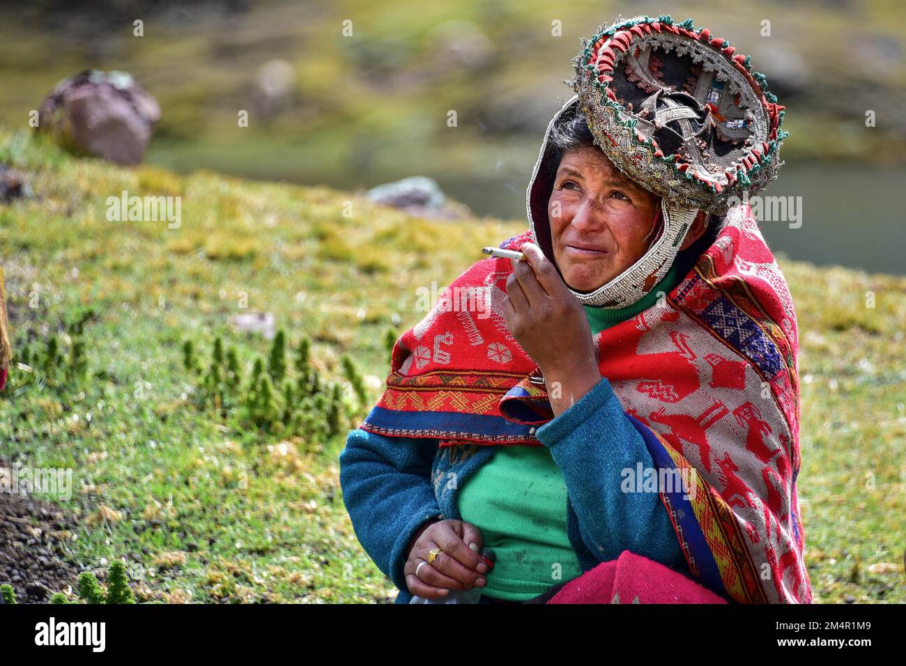 Quechua-Indianerin in traditionellen Kleidern sitzt auf einer Wiese und raucht bei der Pacha-Mama-Zeremonie, Anden, Ollantaytambo, Urubamba-Tal, in der Nähe von Cusco Stockfoto