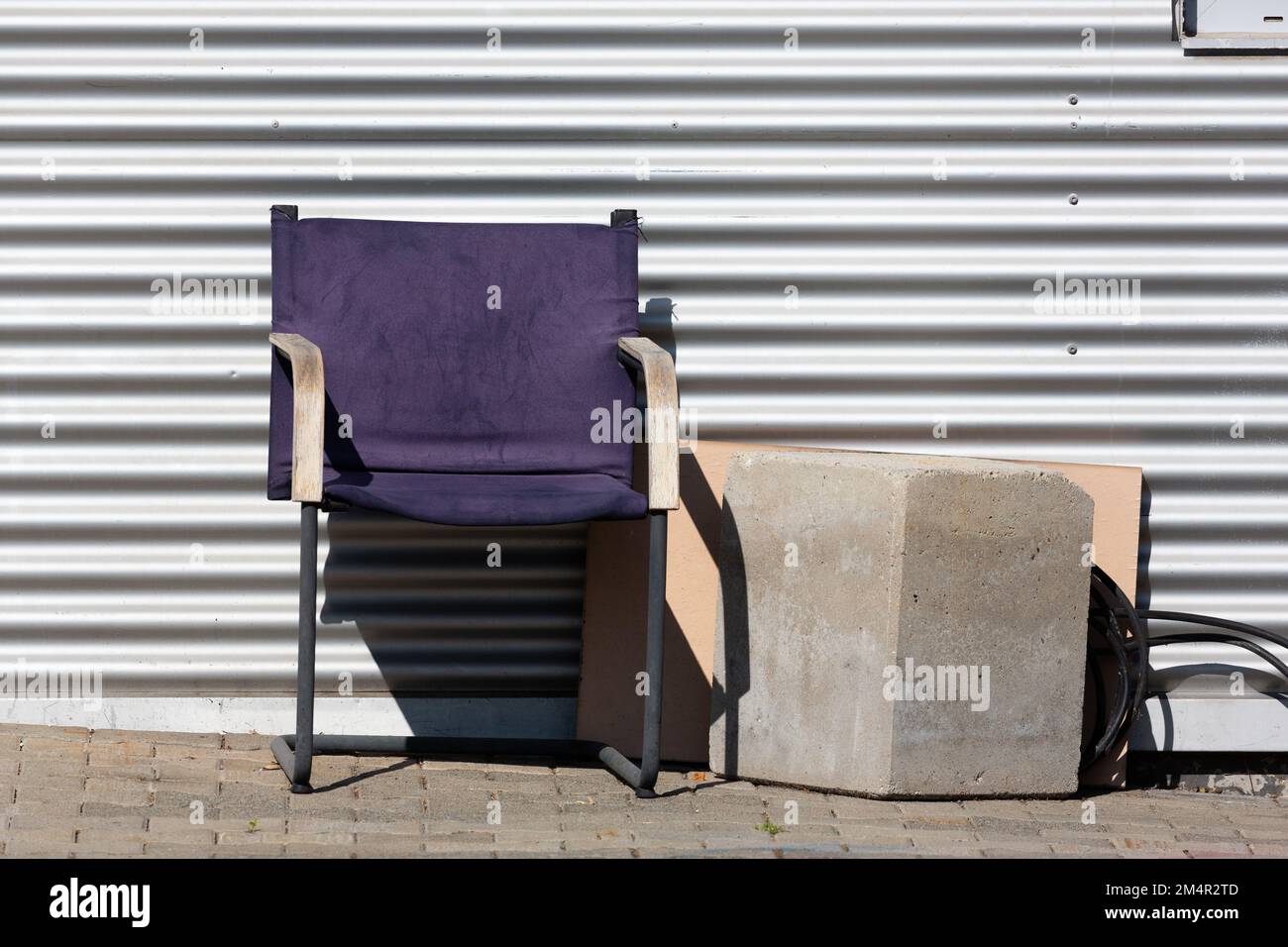 Ein verwitterter Stuhl mit violettem Stoff steht in der Sonne vor einer gerillten Metallwand neben einem Betonblock und lädt Sie ein, sich auszuruhen. Stockfoto