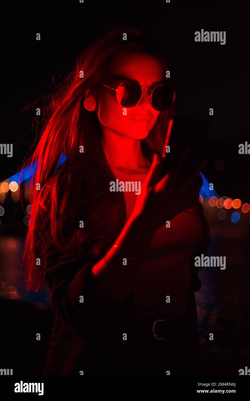 Modische junge Geschäftsfrau, die im Freien eine Sonnenbrille mit einem Mobiltelefon trägt und nachts ein leuchtendes rotes Neonlicht trägt Stockfoto