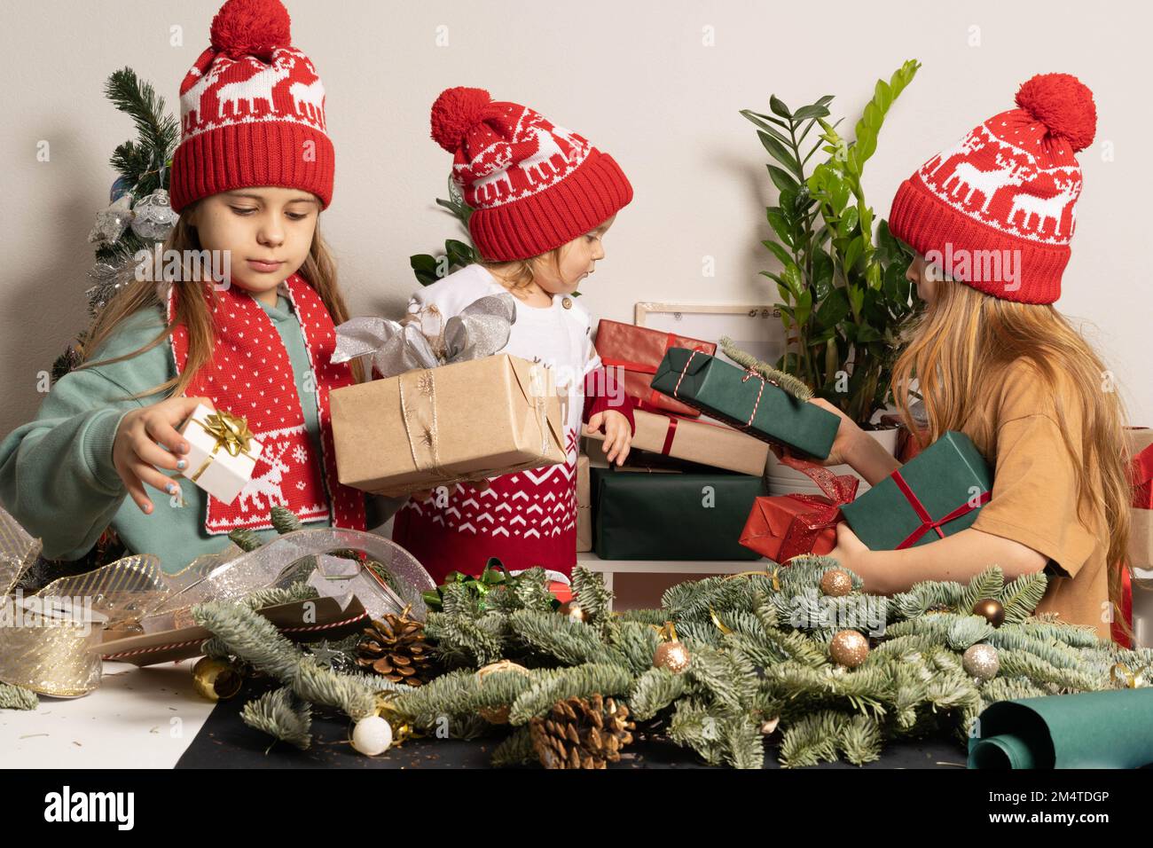 Drei Kinder in roten Weihnachtshüten packen Geschenke und machen Weihnachtsdekorationen. Das Konzept des Hobbys, der Familie, des Urlaubs und des umweltfreundlichen Lebensstils. Stockfoto