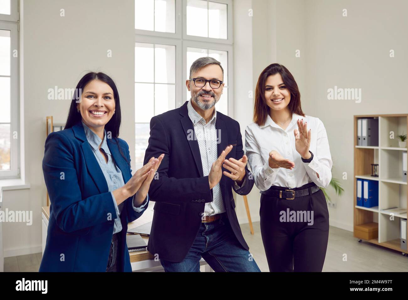 Brutaler, grauhaariger, glücklicher Mann und zwei schöne, lächelnde junge Geschäftsfrauen applaudieren, wenn sie in die Kamera schauen. Stockfoto