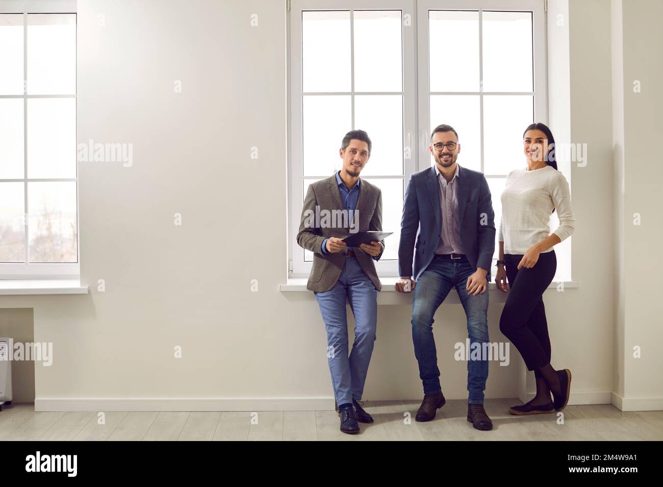 Ein Team aus drei glücklichen, lächelnden jungen Geschäftsleuten, die an einem Bürofenster zusammenstehen Stockfoto