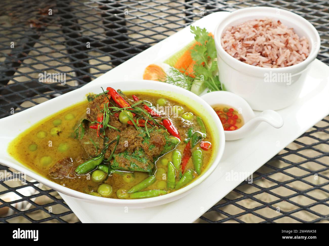 Warmes und würziges grünes thailändisches Curry mit Rindfleisch und eine Schüssel gekochten roten Thai Cargo Reis im Hintergrund Stockfoto