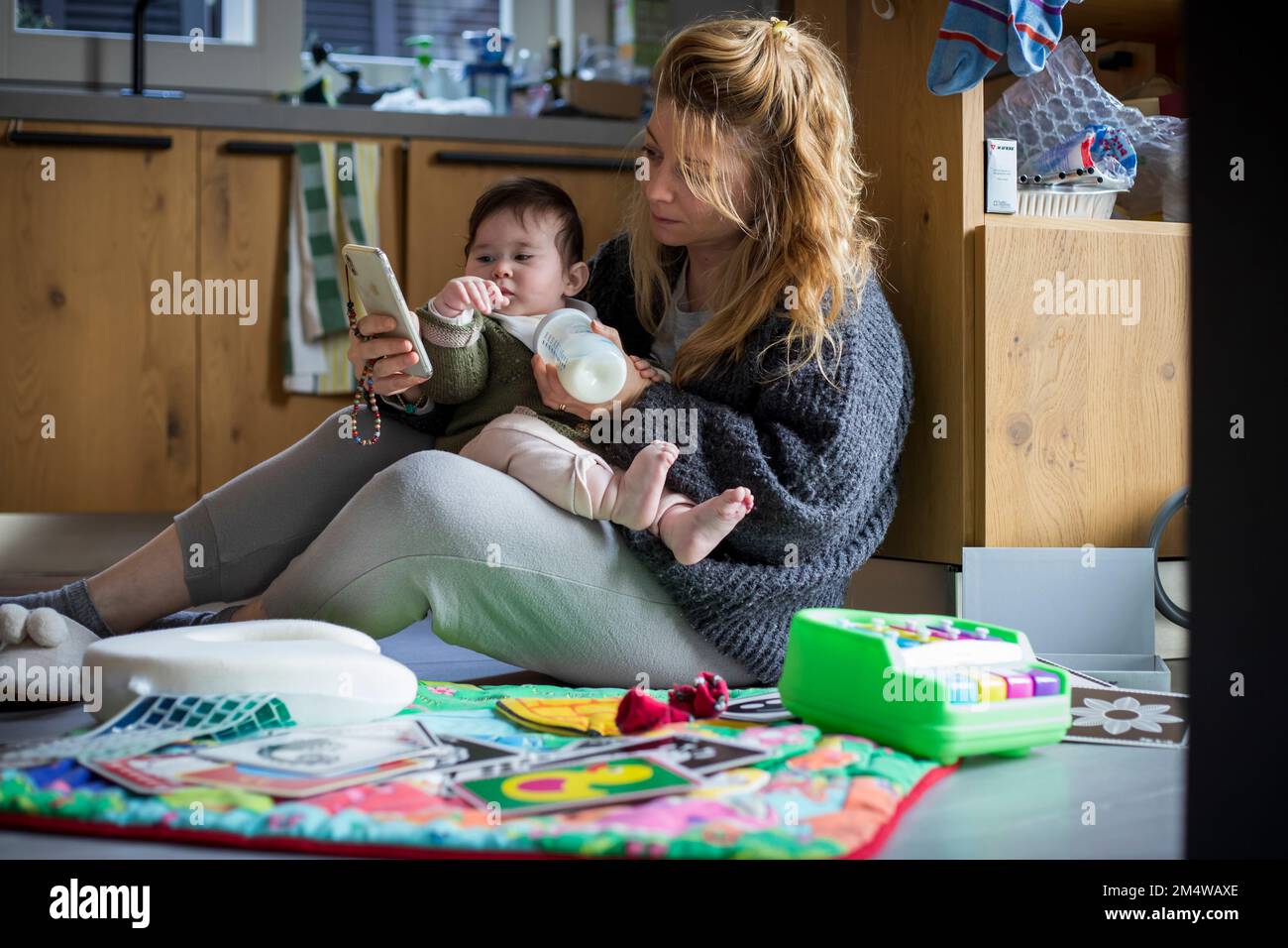 Mutter und Baby spielen morgens in der Küche des chaotischen Hauses mit ihren Hauskleidung und schauen sich die Benachrichtigungen am Telefon an.Alltag Stockfoto