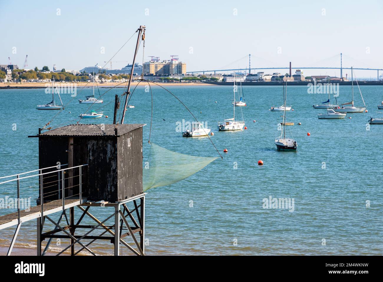 An einem sonnigen Sommertag steht eine hölzerne Angelkabine mit einem großen quadratischen Aufzugsnetz namens „carrelet“ am Strand gegenüber der Saint-Nazaire-Brücke. Stockfoto