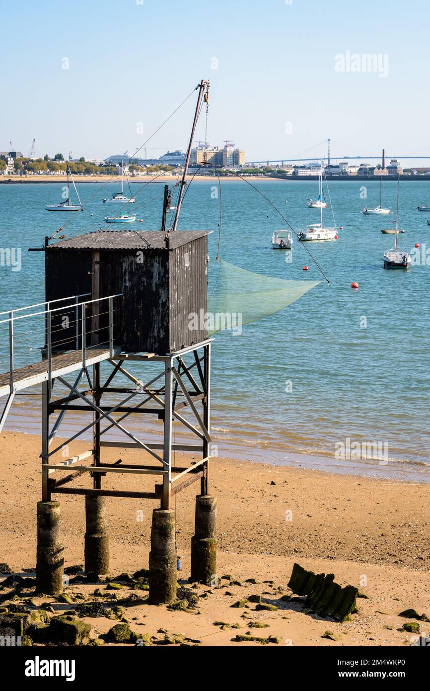 An einem sonnigen Sommertag steht eine hölzerne Angelkabine mit einem großen quadratischen Aufzugsnetz namens „carrelet“ am Strand gegenüber der Saint-Nazaire-Brücke. Stockfoto