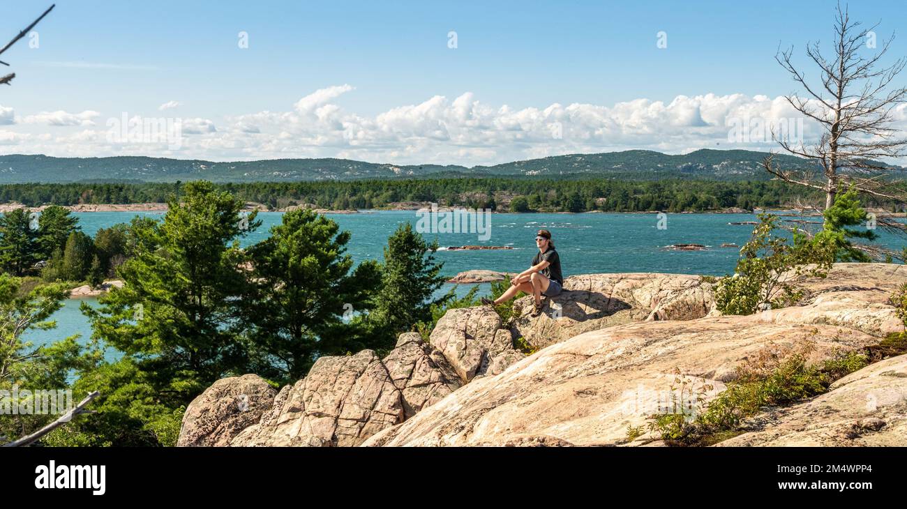Junger Mann sitzt auf dem Solomon Island Bluff, mit den La Cloche Mountains im Hintergrund. Stockfoto
