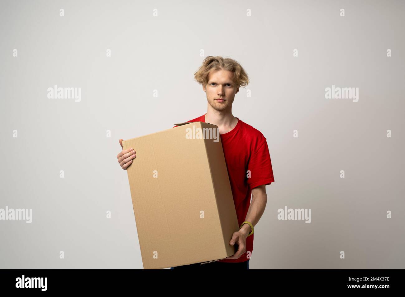 Der Angestellte in roter Uniform arbeitet als Kurier des Händlers und hält einen leeren Karton isoliert auf weißem Hintergrund im Studio. Konzept des Lieferservice Stockfoto