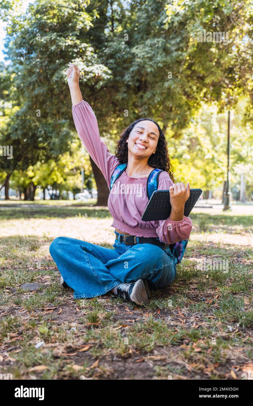 Eine Studentin aus Lateinamerika feiert und hält ein Tablet in der Hand, während sie draußen in einem Park auf dem Rasen sitzt Stockfoto