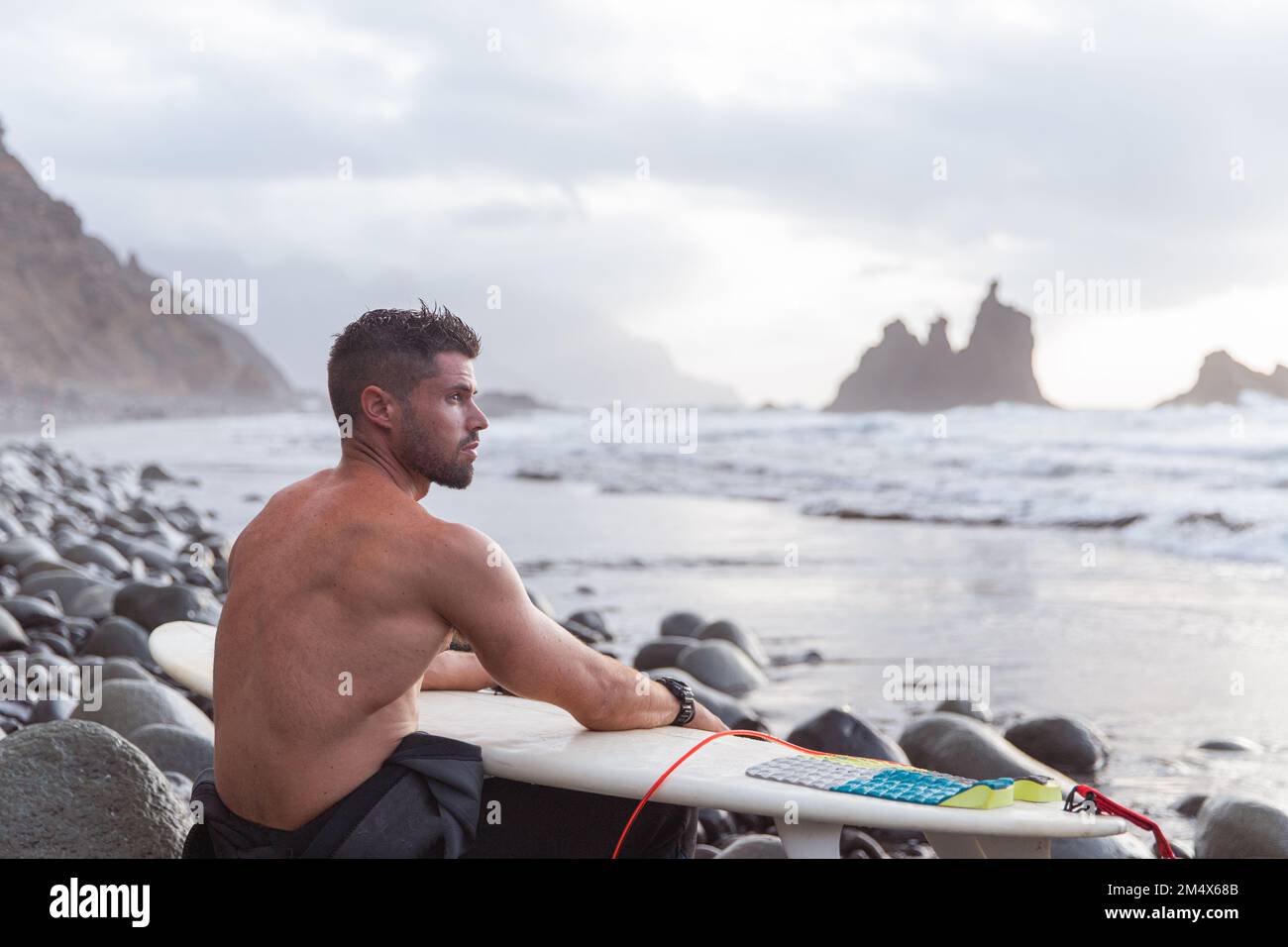 Surfer sitzt am Strand mit seinem Surfbrett, sportlicher Typ, macht Extremsport, Foto mit Kopierraum Stockfoto