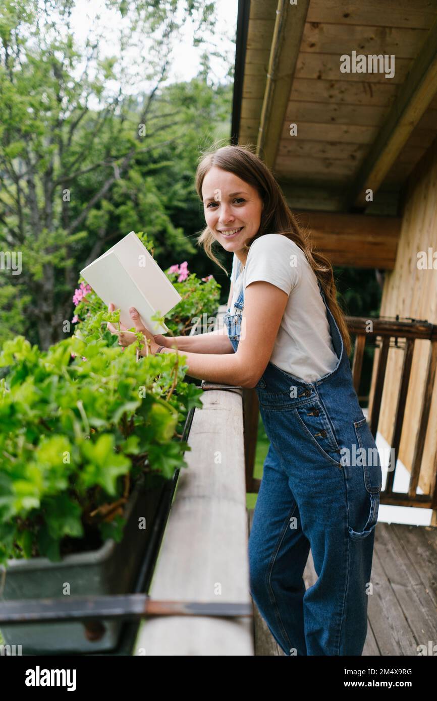 Lächelnde Frau mit Buch, das auf dem Geländer auf dem Balkon liegt Stockfoto