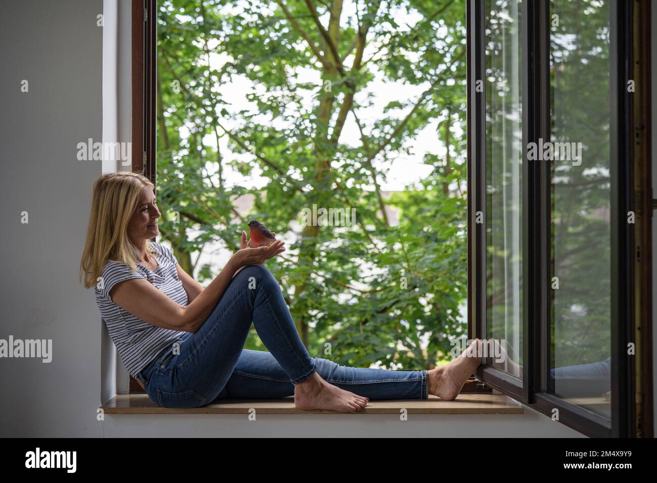 Eine reife Frau, die einen Vogel hält und zu Hause am Fenster sitzt Stockfoto