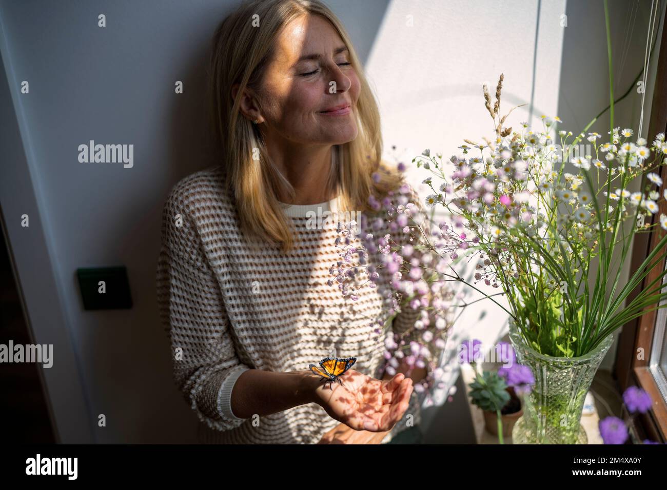 Reife Frau, die zu Hause mit Schmetterling und Sonnenlicht über dem Gesicht steht Stockfoto