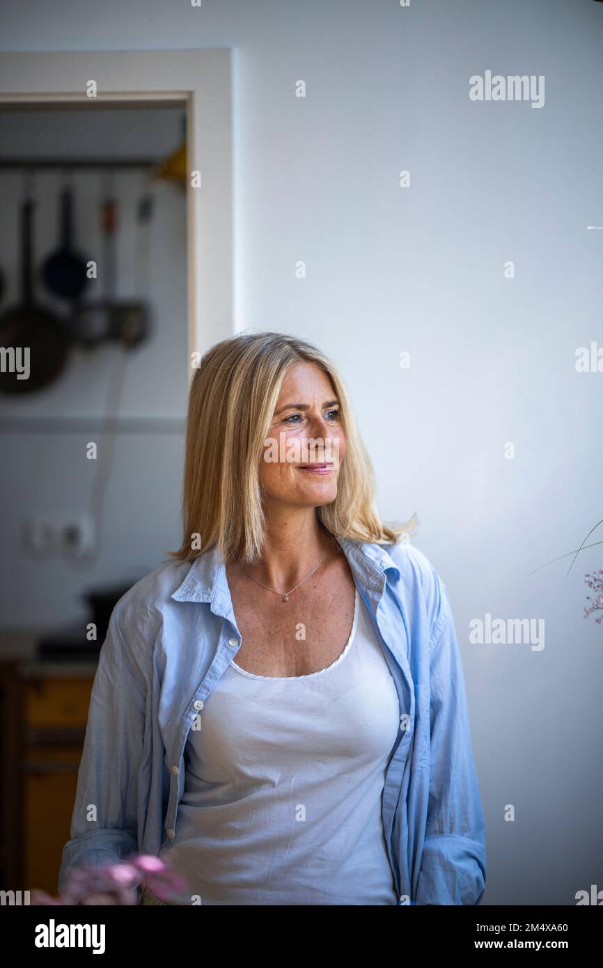 Kontemplative, reife Frau, die zu Hause mit einem vollständig aufgeknöpften Hemd steht Stockfoto