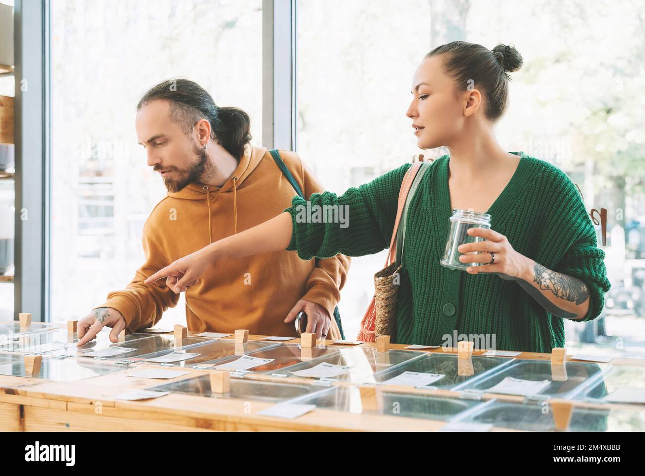 Eine Frau mit einem Glas, das auf den Tresen zeigt und mit einem Mann im Lebensmittelladen spricht Stockfoto