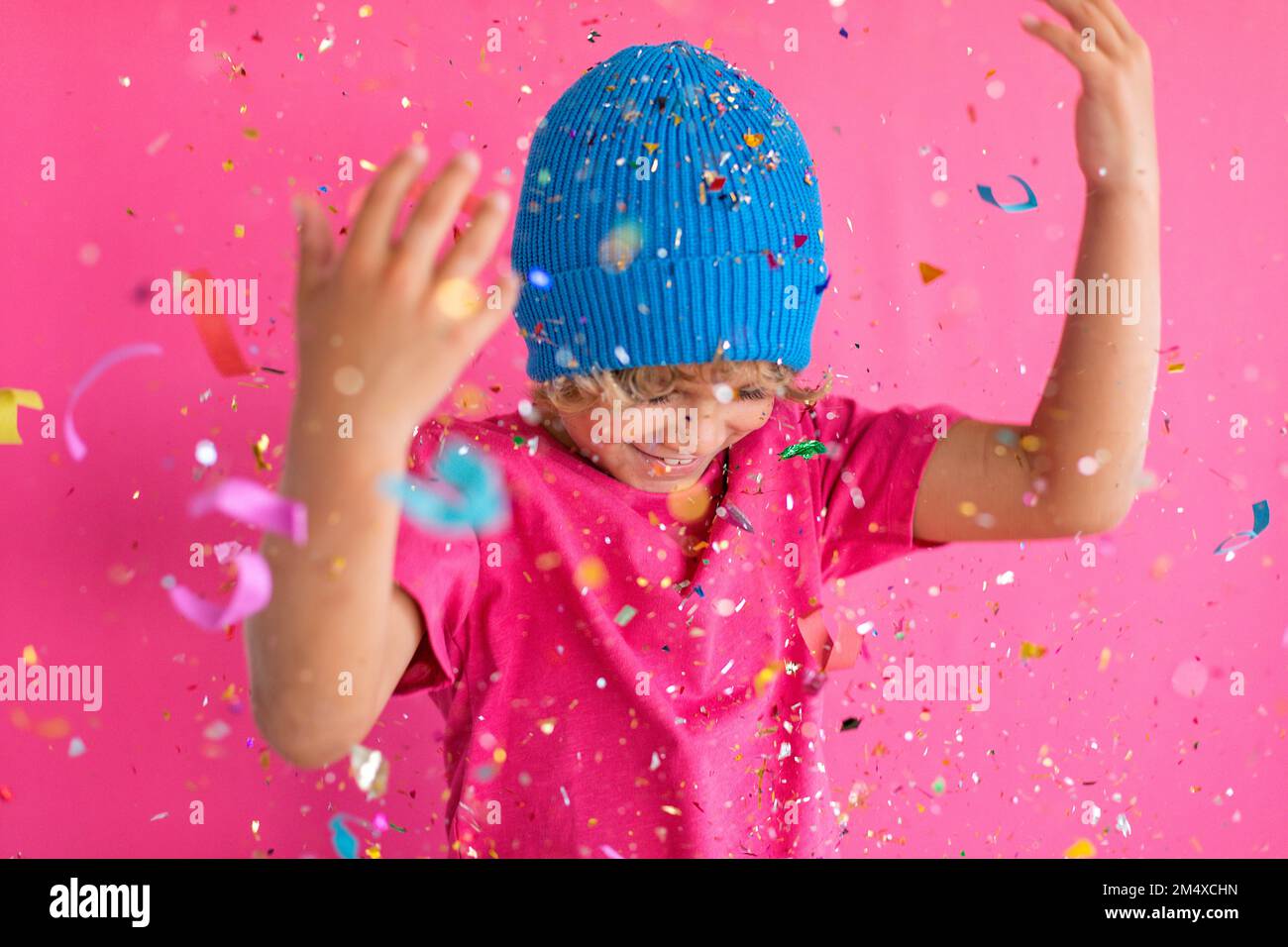 Glücklicher Junge mit blauer Strickmütze, der mit Konfetti vor pinkfarbenem Hintergrund spielt Stockfoto