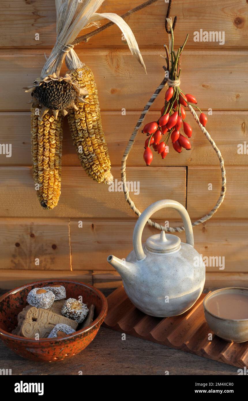 Rosenhüften, Maiskolben, Keramikteekanne und Schüssel mit Keksen und Früchtekuchen Stockfoto