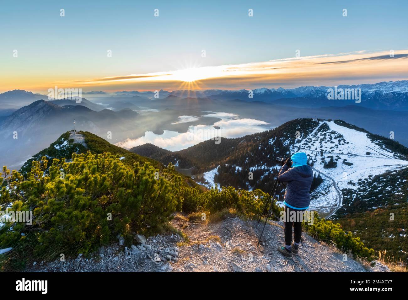 Deutschland, Bayern, Frau, die die umliegende Landschaft bei Sonnenaufgang vom Gipfel des Herzogstands fotografiert Stockfoto