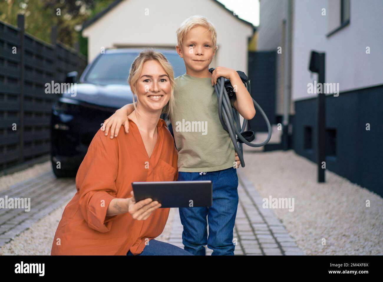 Mutter mit Tablet-PC, Sohn, der den Stecker des Ladegeräts für Elektrofahrzeuge im Vorgarten hält Stockfoto