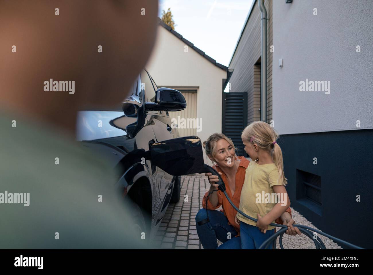 Ein Mädchen sieht sich die Mutter an, die den Stecker für das Ladegerät für Elektrofahrzeuge im Vorgarten hält Stockfoto