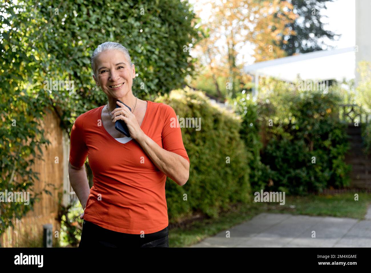 Lächelnde Seniorin mit orangefarbenem T-Shirt und Handy im Garten Stockfoto