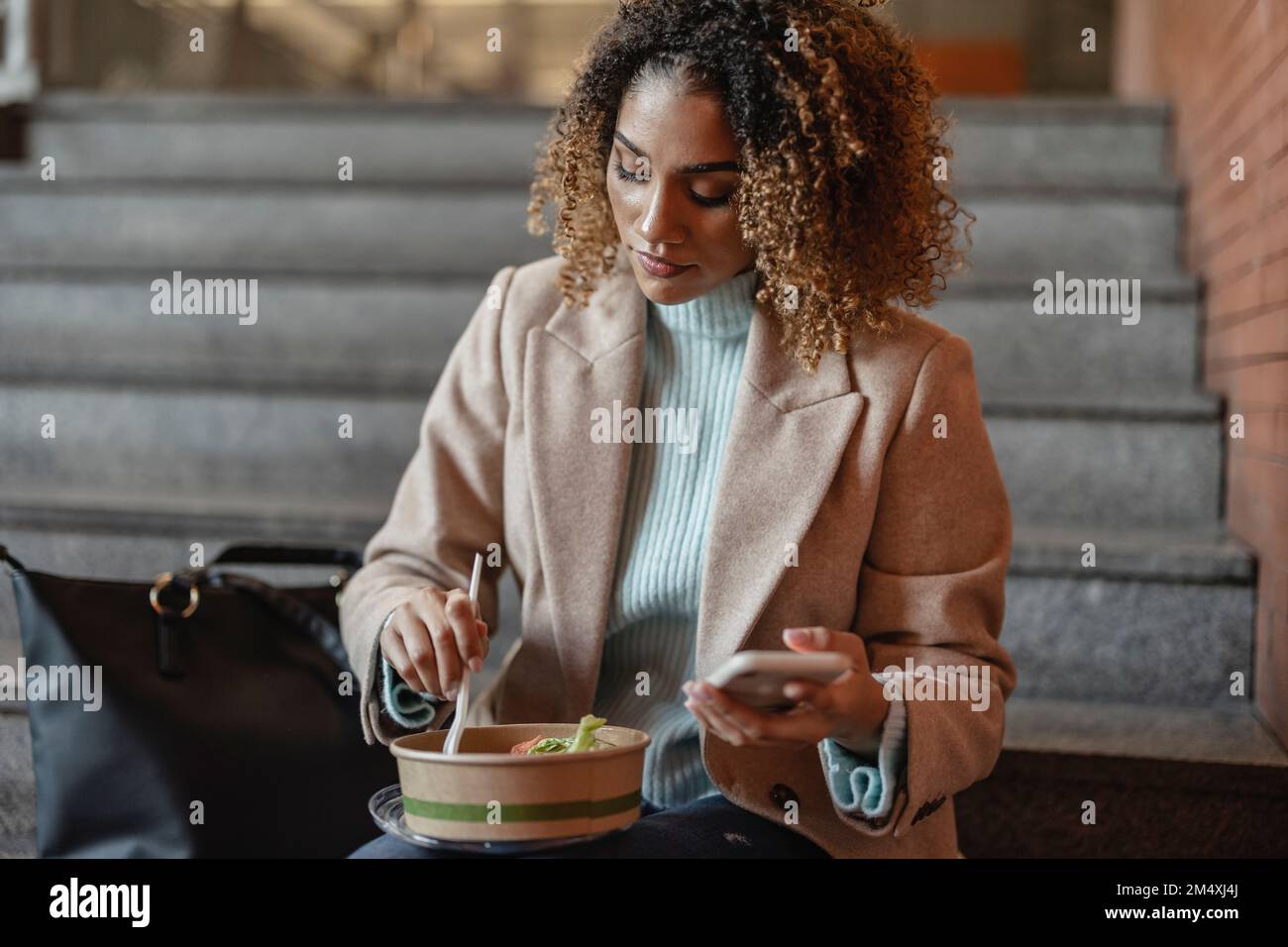 Junge Frau, die ein Handy hält und Essen auf der Treppe hat Stockfoto