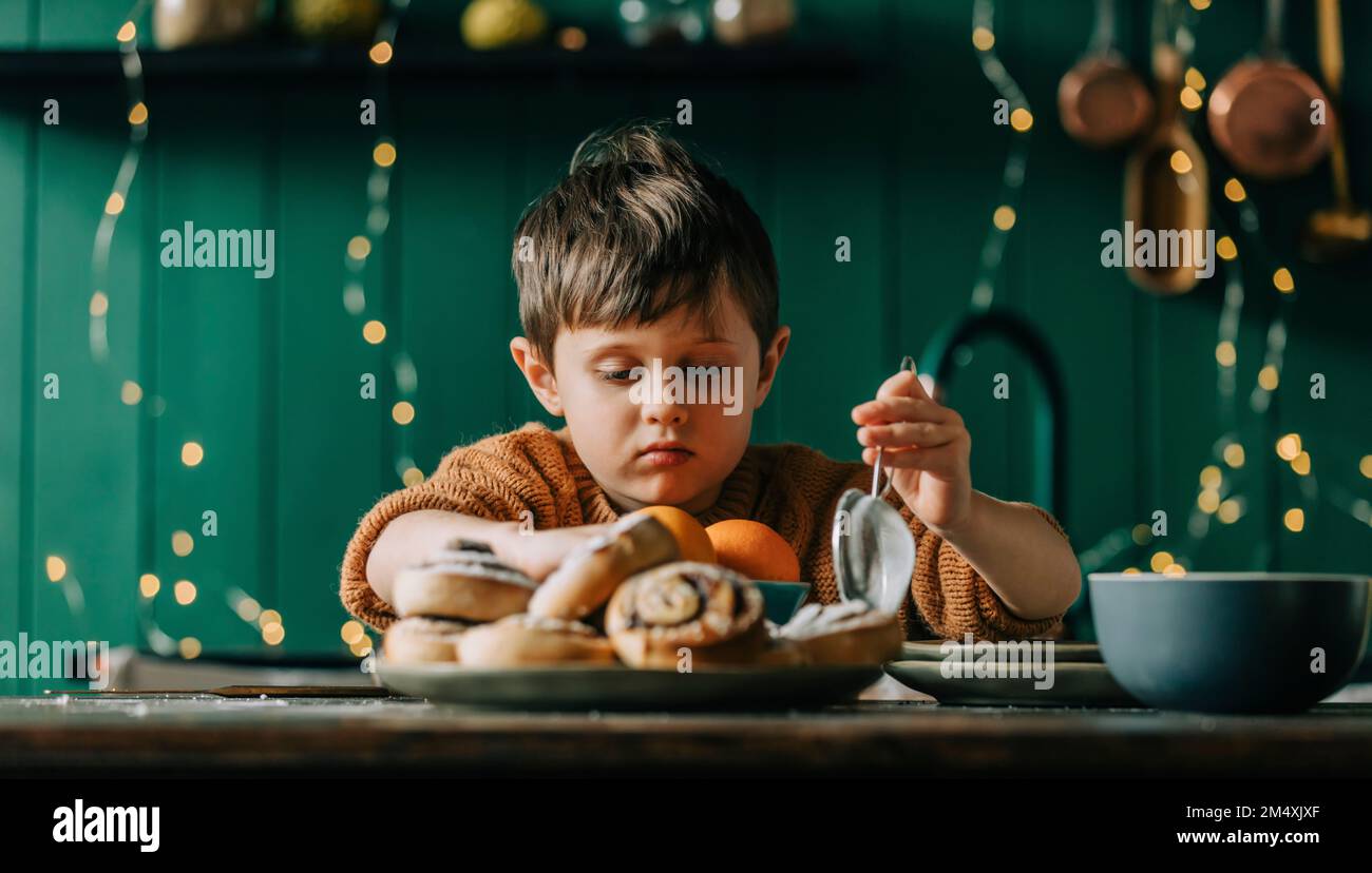 Ein Junge, der ein Sieb vor Zimtbrötchen und Orangen auf dem Tisch hält Stockfoto