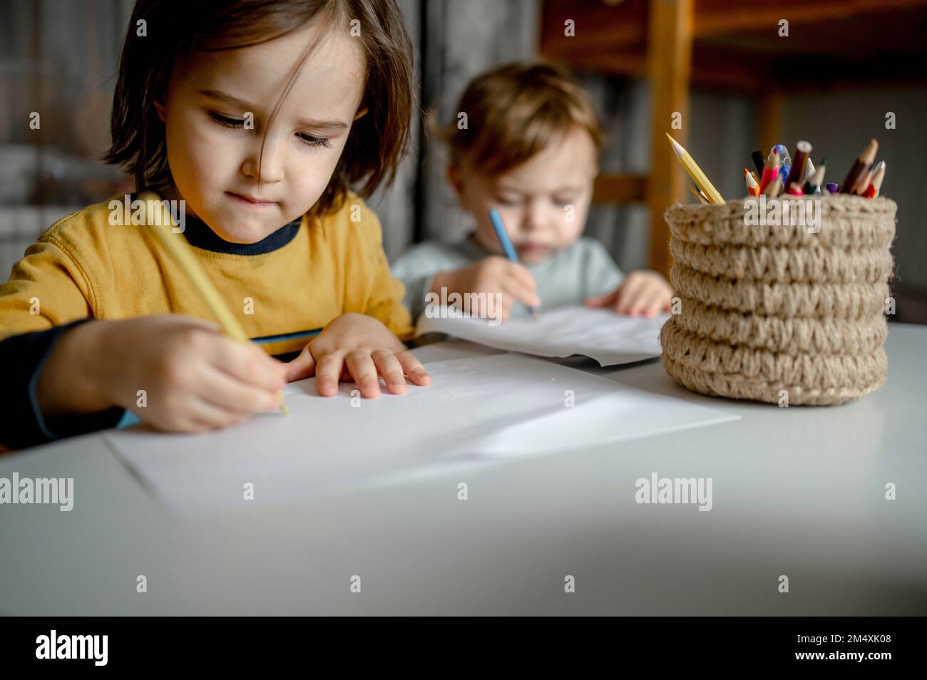 Ein Junge mit einem Bruder, der auf Papier mit bunten Bleistiften in einem Jutebehälter malt Stockfoto