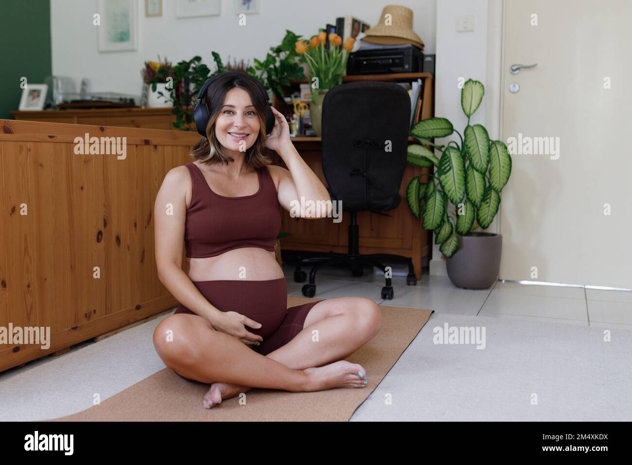Glückliche Schwangere, die Musik über kabellose Kopfhörer hört, die zu Hause auf einer Yoga-Matte sitzen Stockfoto