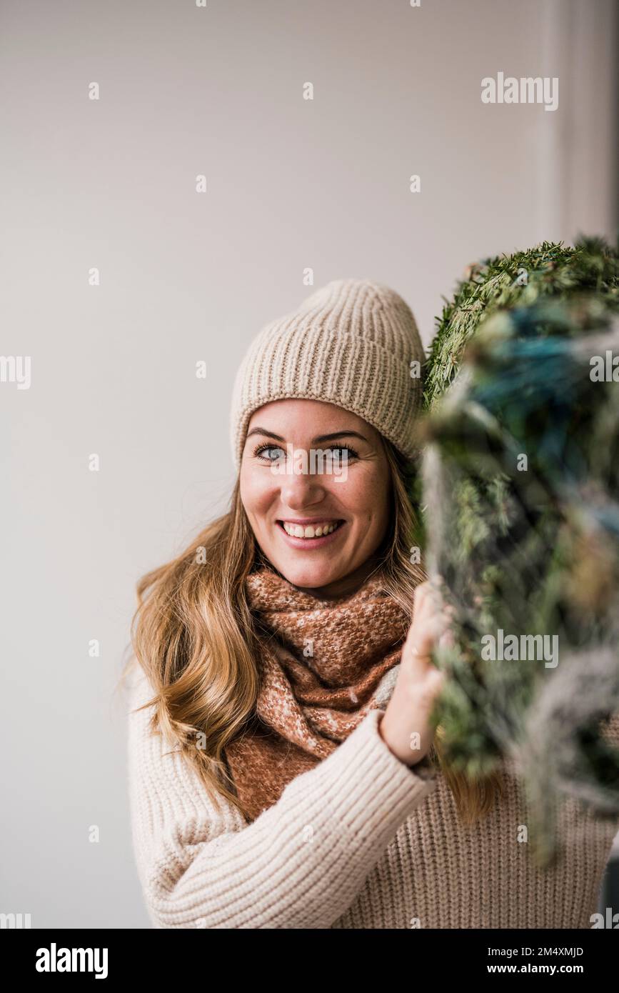 Lächelnde Frau, die zu Hause einen Weihnachtsbaum auf der Schulter trägt Stockfoto