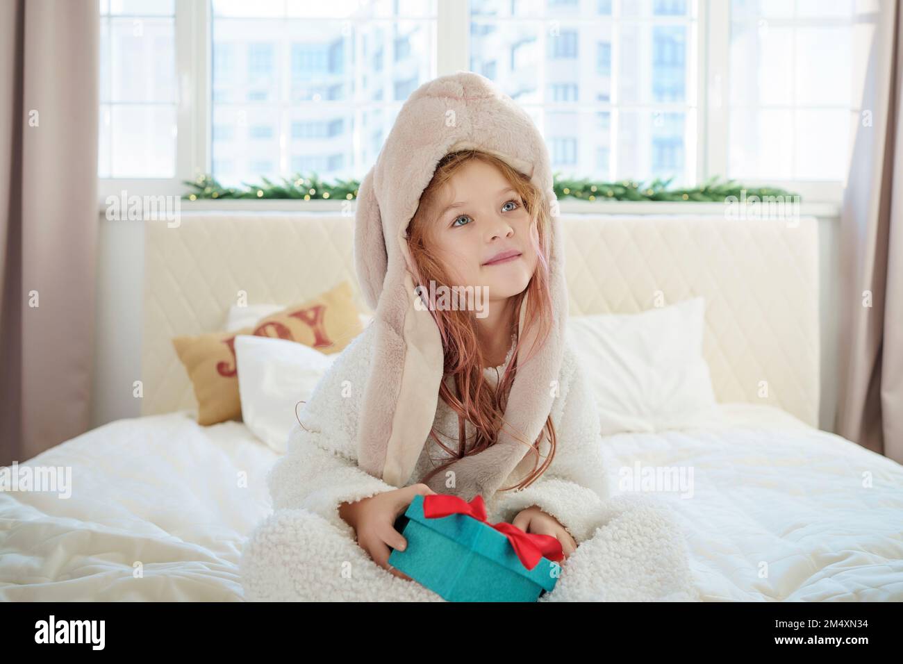 Ein Mädchen mit Hut, das mit einem Weihnachtsgeschenk auf dem Bett sitzt und zu Hause nachdenkt Stockfoto
