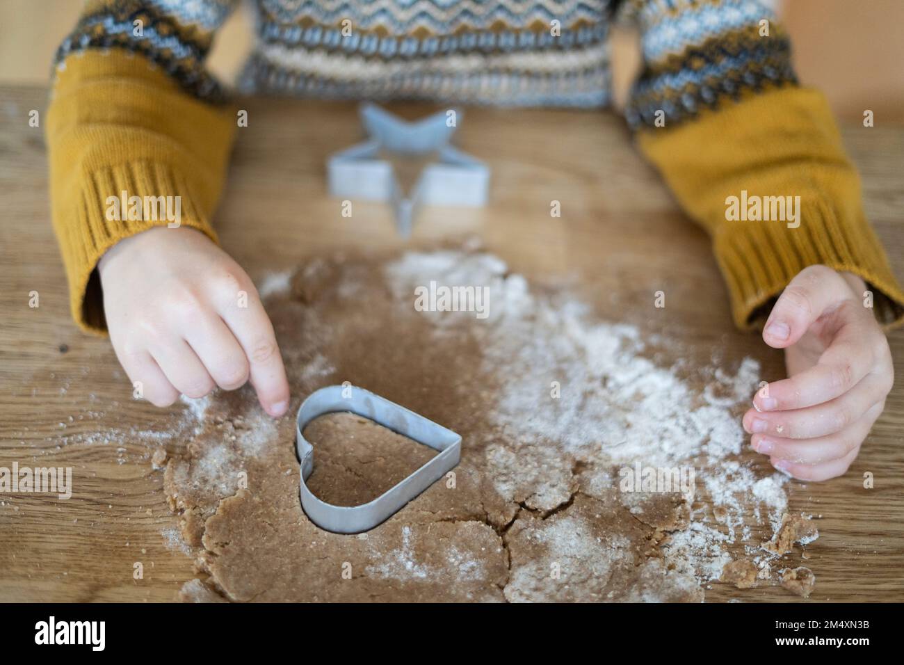 Hände eines Jungen, der Kekse mit herzförmigem Messer auf dem Tisch ausschneidet Stockfoto