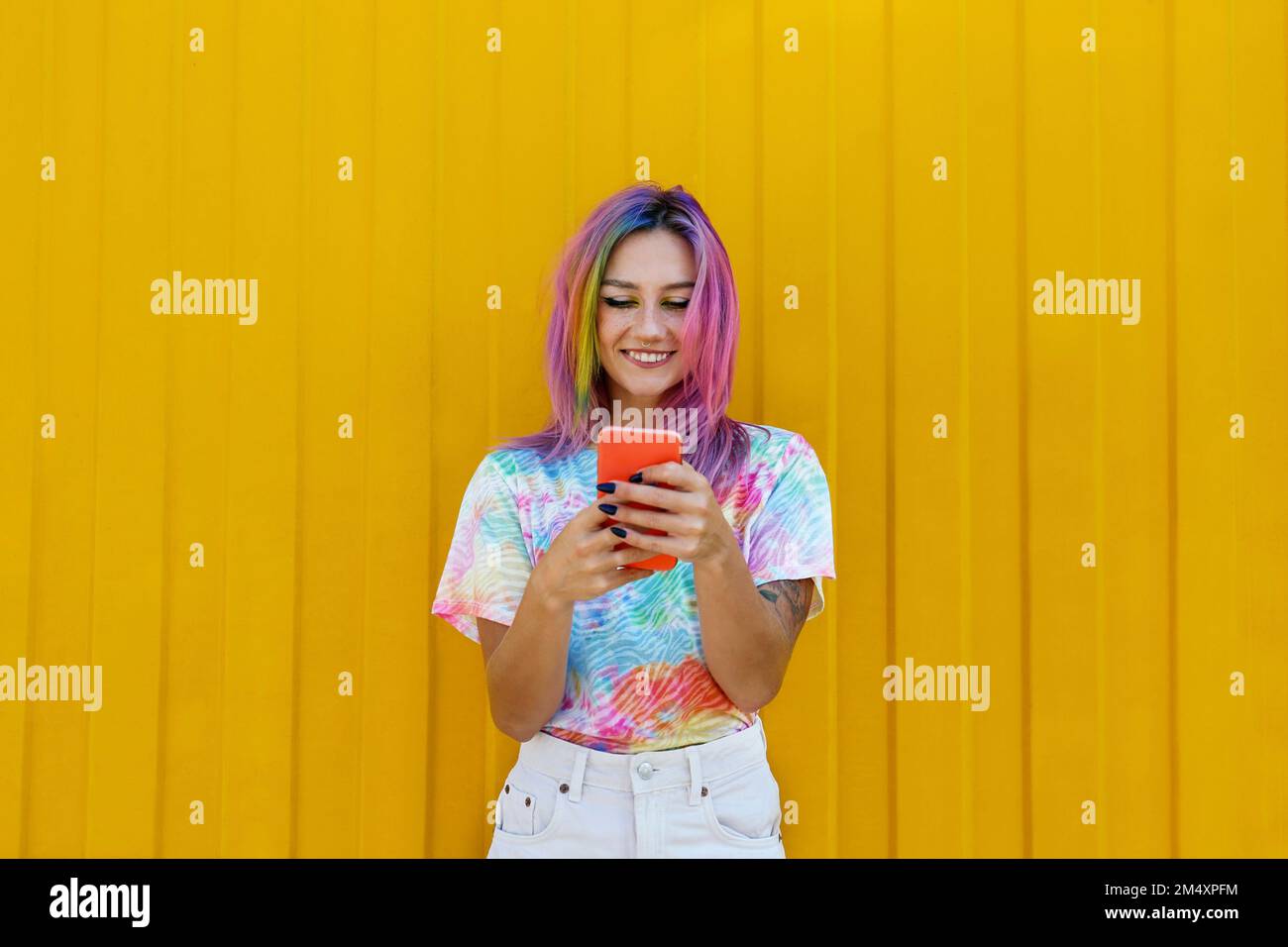 Lächelnde junge Frau, die ein T-Shirt mit Krawatte und Smartphone vor der gelben Wand trägt Stockfoto