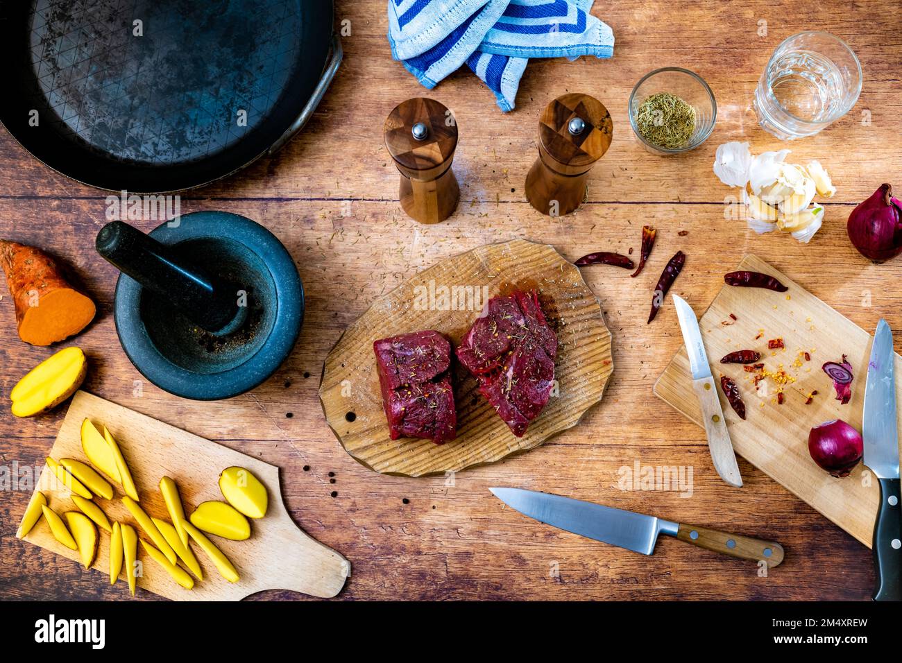 Beschreibung: Gewürzfiletsteak aus rohem Rindfleisch auf einem Holzbrett mit Stahlpfanne, Mörtel, Küchenhandtuchsalz und Pfefferstreuer und rohen Kartoffelspalten Stockfoto