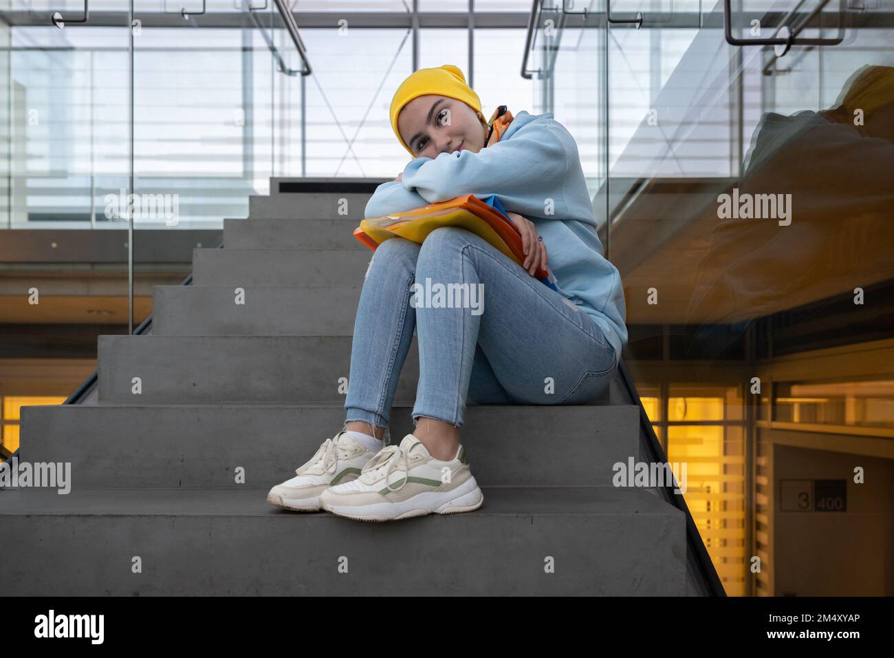 Ein Mädchen mit farbigen Ordnern in der Hand sitzt auf einer Steintreppe zwischen den Etagen und legt den Kopf auf die Knie Stockfoto