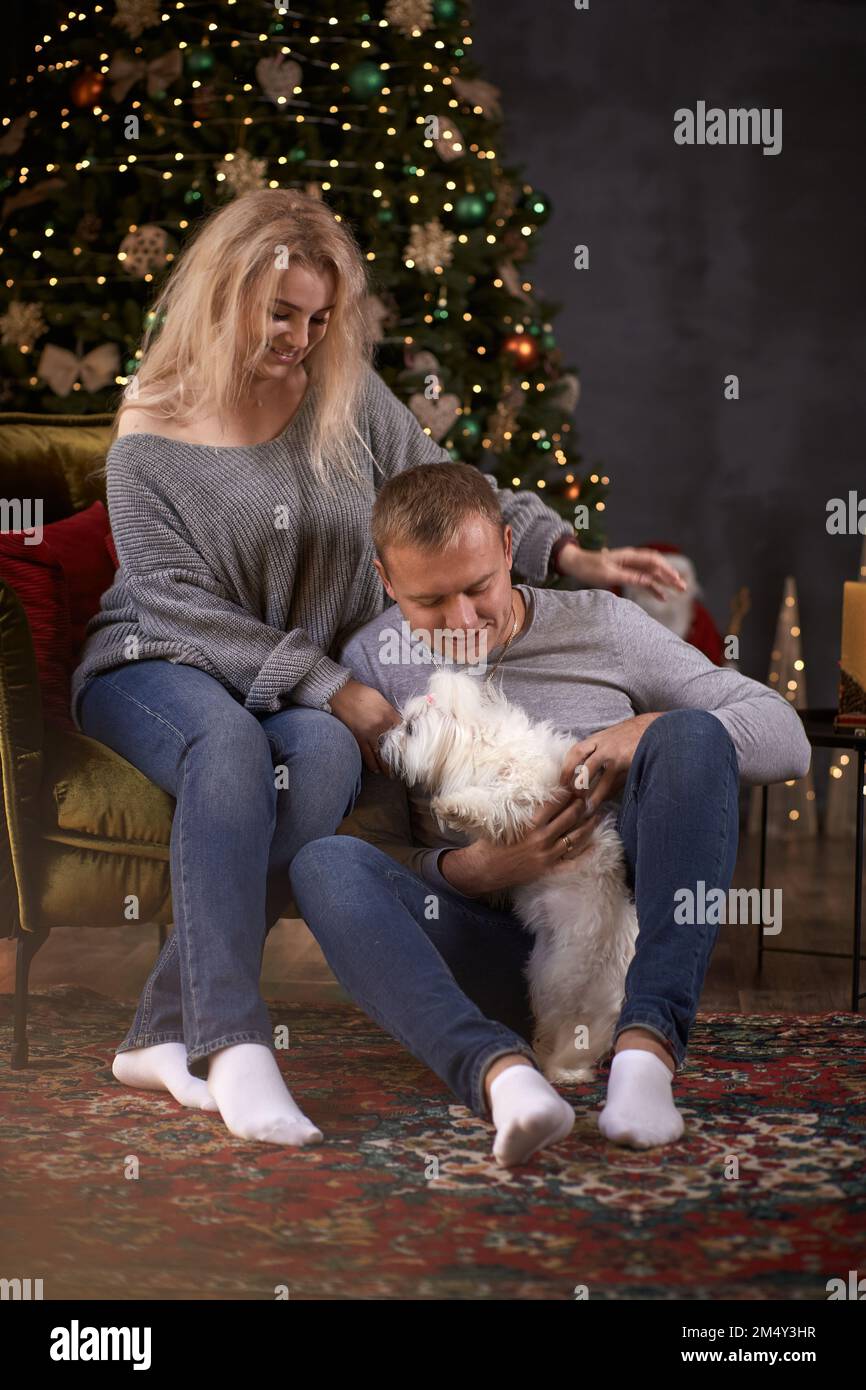 Ein romantisches Paar mit seinem Hund macht sich in der Nähe des Weihnachtsbaums in festlicher, ästhetischer, gemütlicher Inneneinrichtung lustig. Gute Laune der Weihnachts-Liebesgeschichte. Ein ehrlicher, wahrer Moment Stockfoto