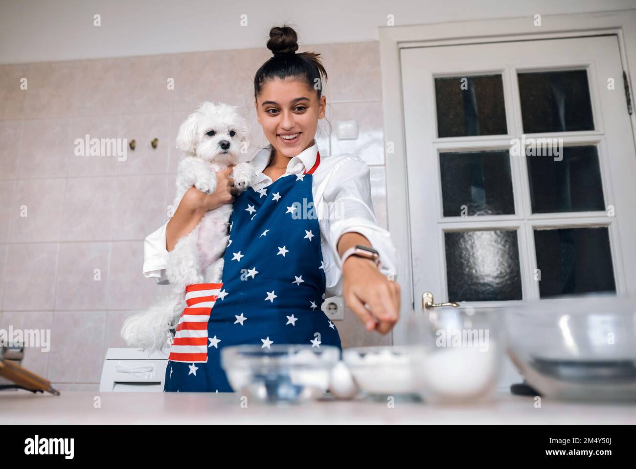 Lächelnde junge Frau zeigt mit dem Finger in die Küche, während sie einen süßen weißen maltesischen Hund hält Stockfoto