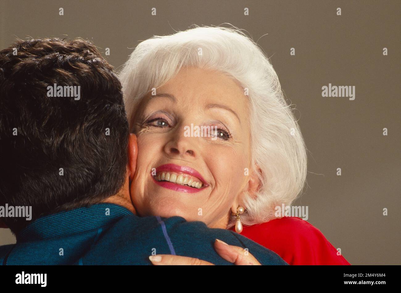 Ältere grauhaarige Weiße, die einen jungen Mann umarmt, sie lächelt und er wird von hinten fotografiert Stockfoto